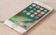 iPhone 7 i hvid ligger på et træbord med forsiden opad. Den er tændt og er på hjemmeskærmen med alle de præinstallerede apps. Man kan ane at det er farven Rosa Guld der er på bagsiden af telefonen.
